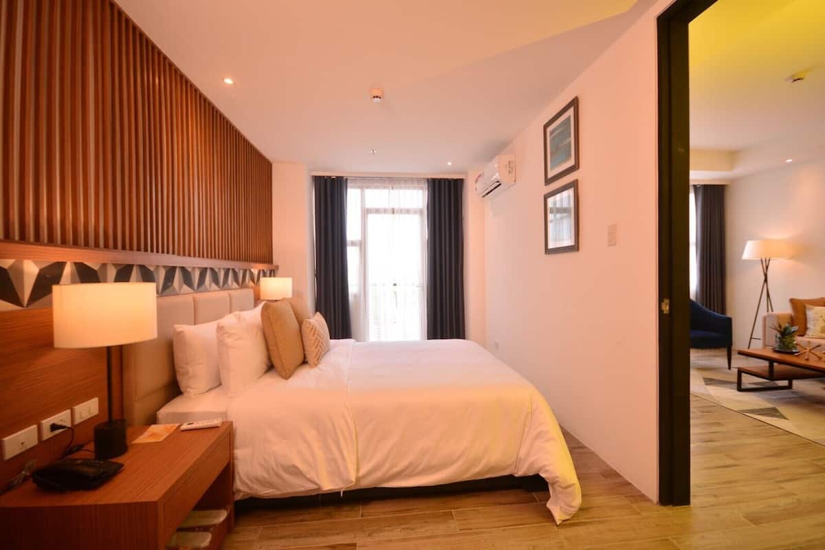 Una habitación de hotel con una cama grande, lámparas de noche, un cabecero de madera, un escritorio con una lámpara y vistas al exterior a través de una ventana.