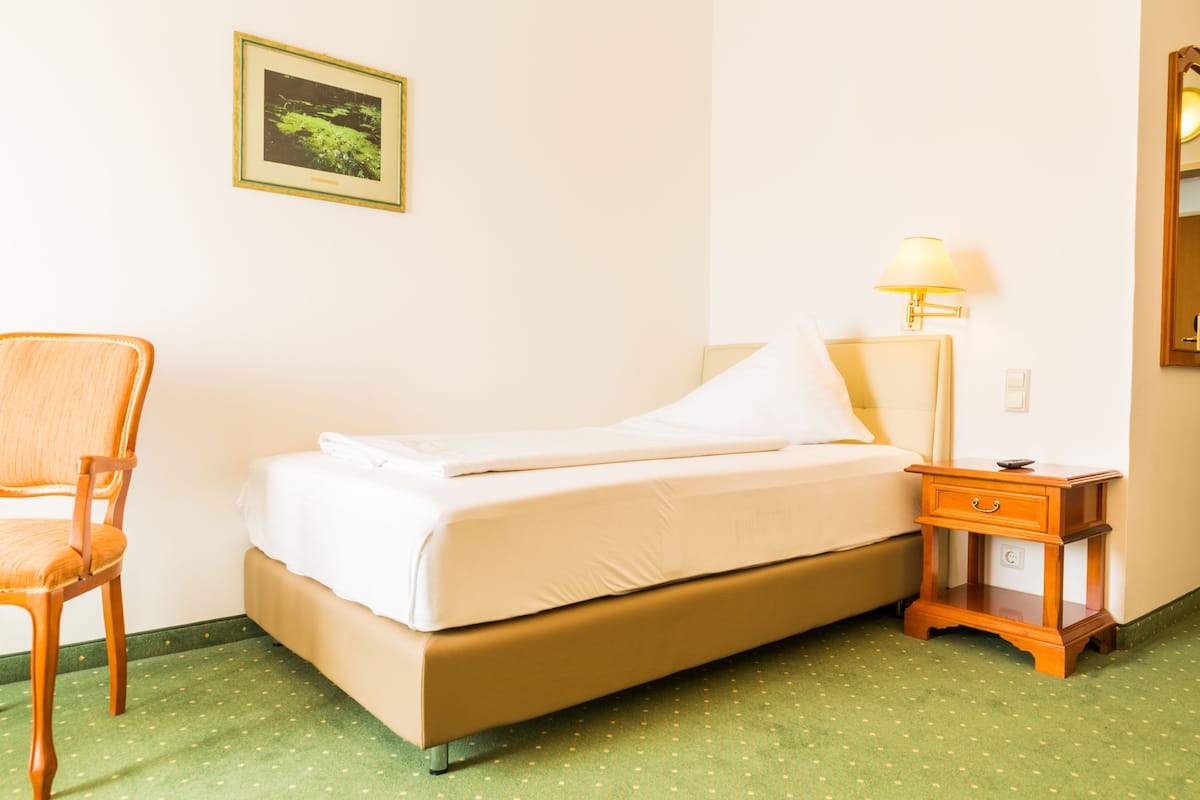 Habitación de hotel con cama, una silla, una mesita de noche con lámpara y un cuadro enmarcado en la pared.