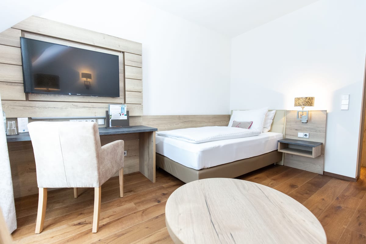 Una habitación de hotel moderna con un escritorio de madera, un televisor de pantalla plana, una cama con sábanas blancas, una mesa redonda de madera y una mesita de noche con una lámpara.