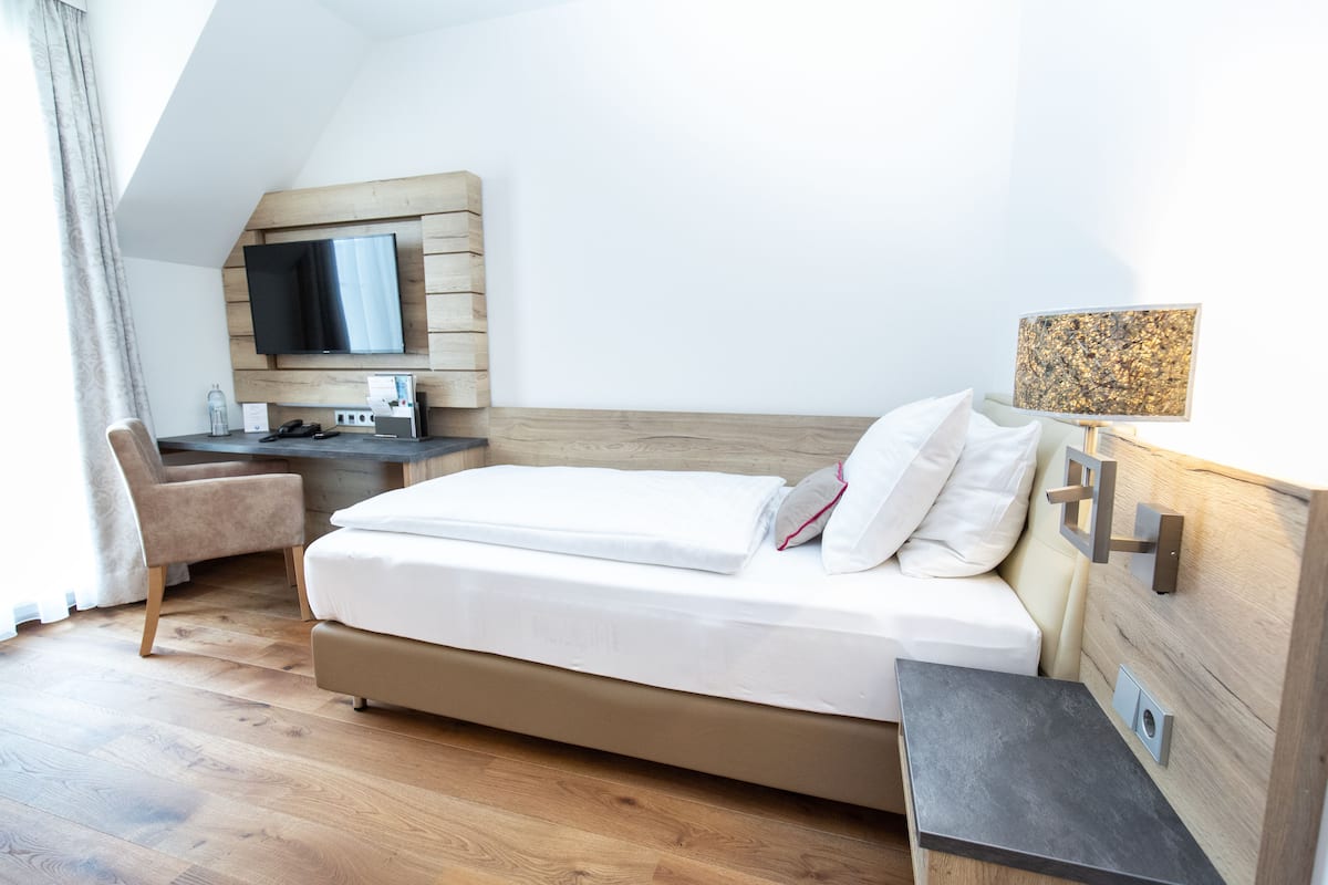 Una habitación de hotel moderna con una cama, un escritorio, una silla, un televisor de pantalla plana montado en un panel de madera y una mesita de noche con una lámpara.
