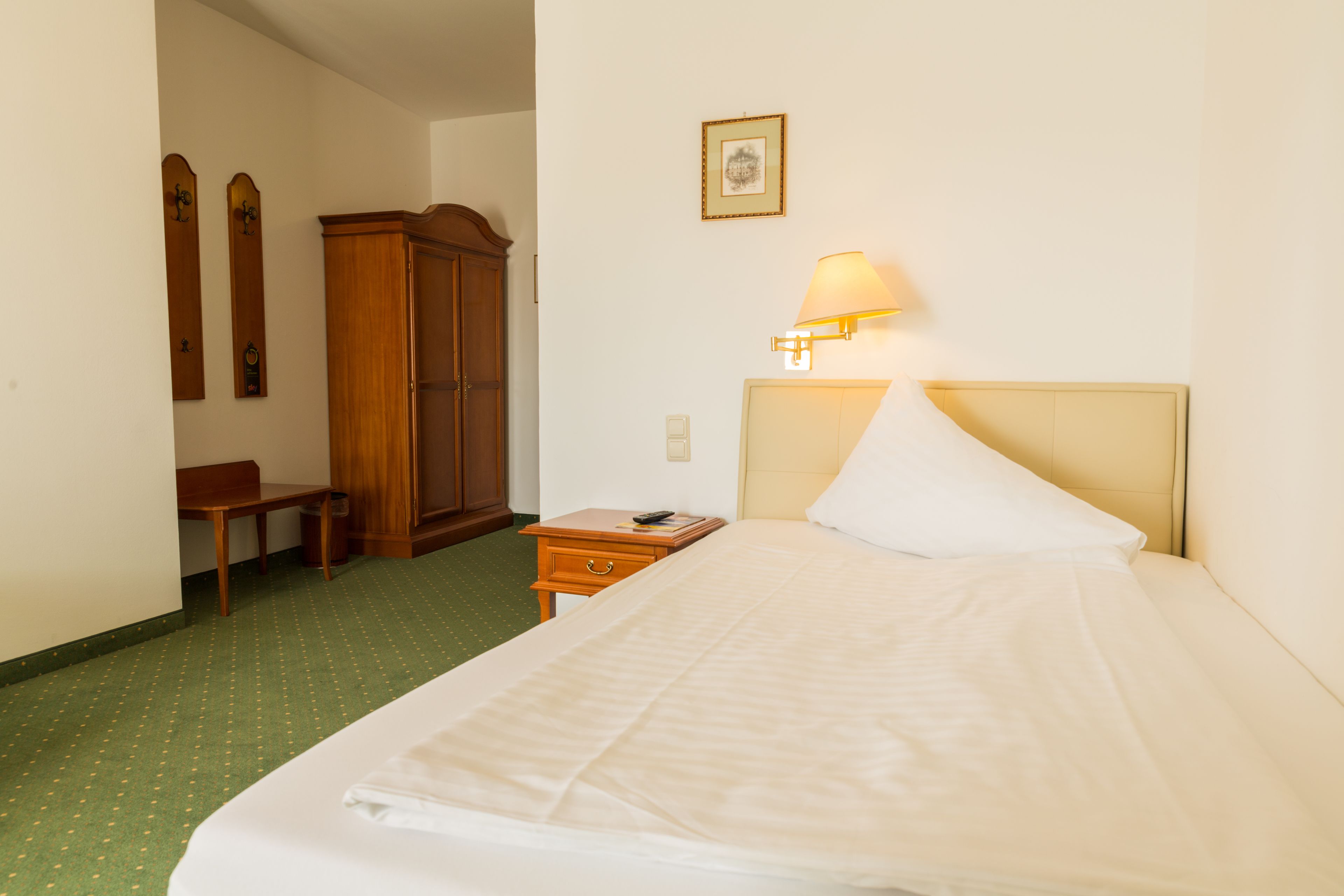 Quarto de hotel com cama, mesa de cabeceira, luminária, guarda-roupa de madeira, cadeira e um quadro na parede.