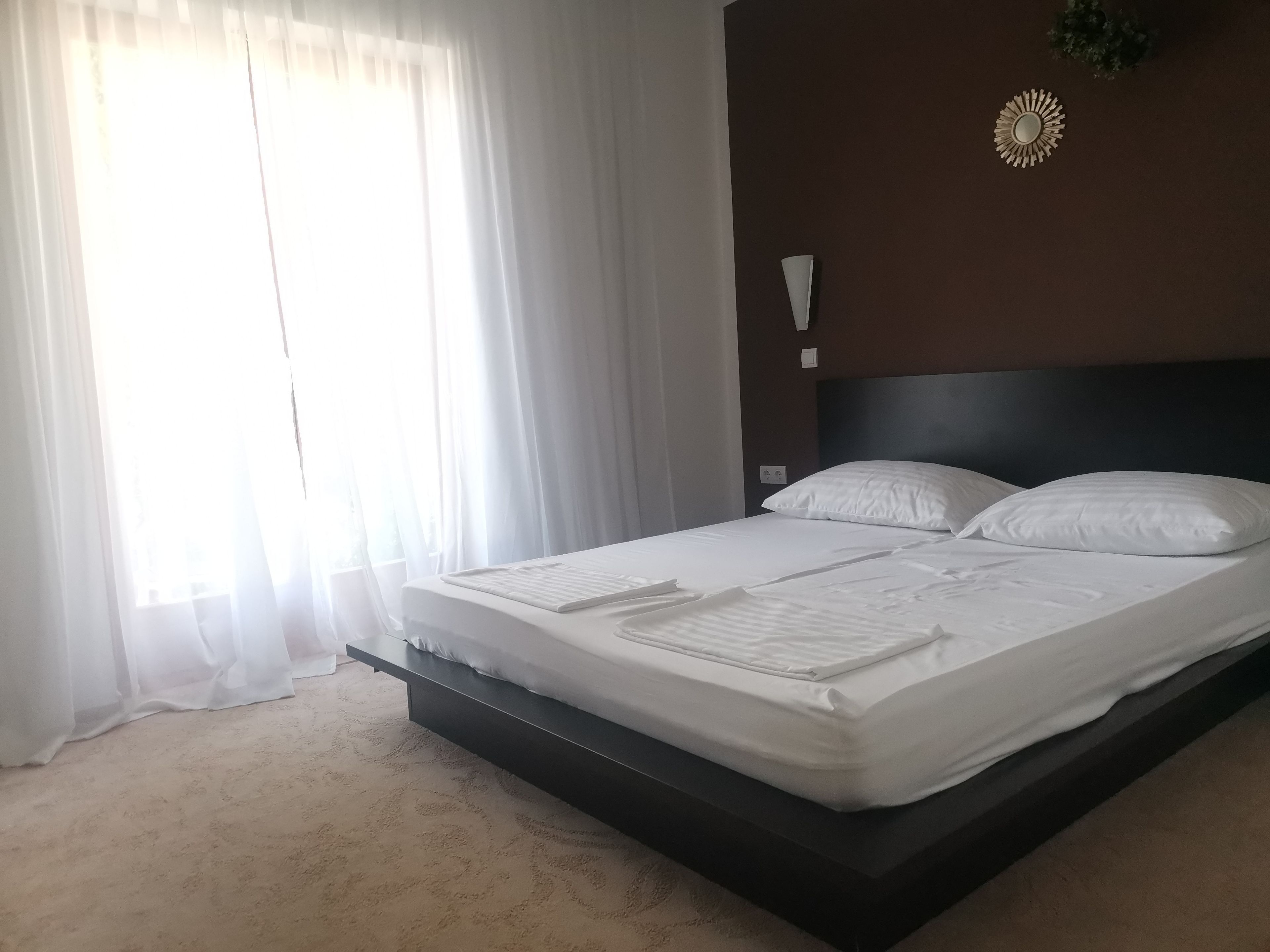 A hotel room with a bed, a nightstand, a wall-mounted light, and sheer curtains.