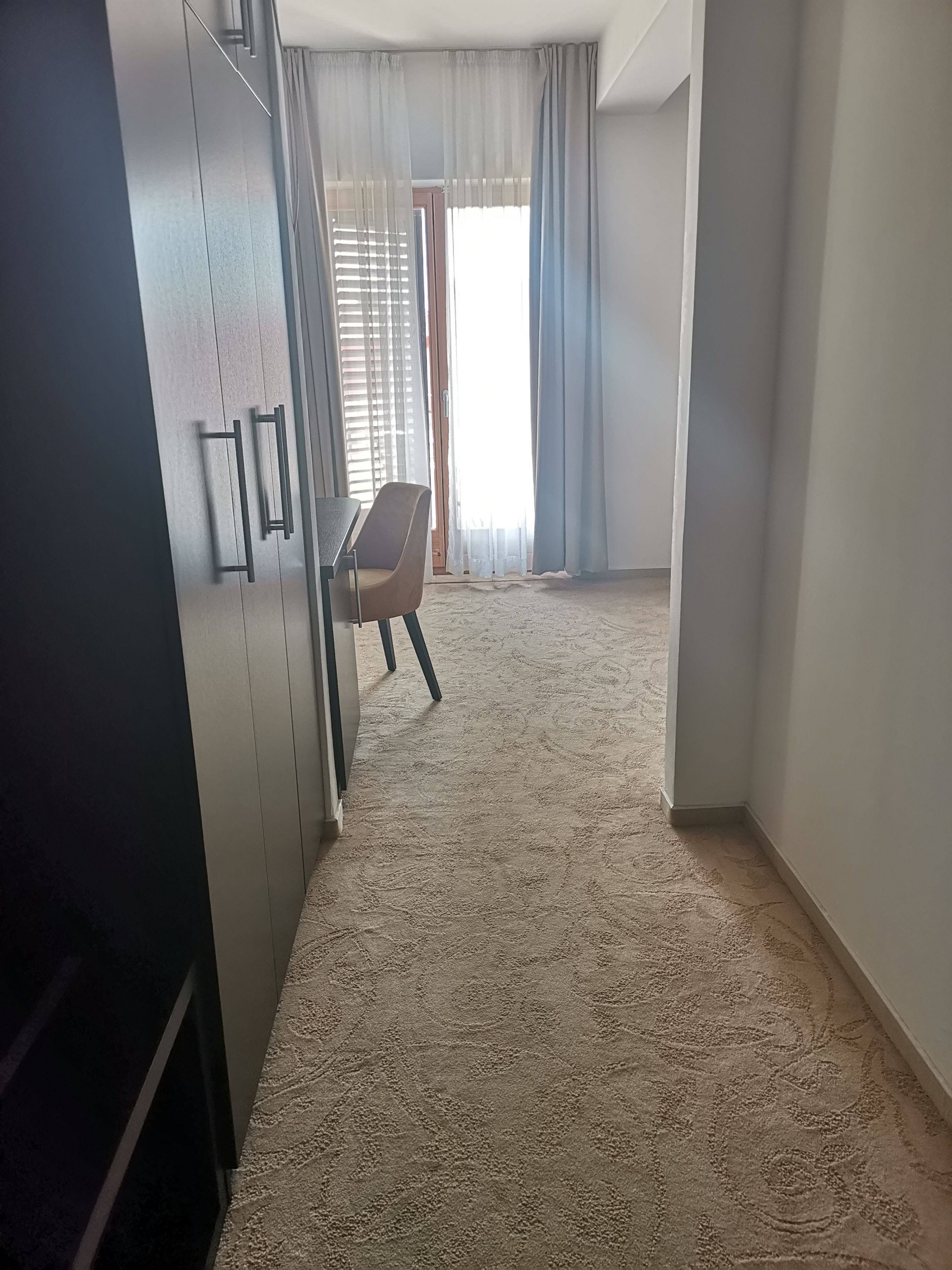 A room with a carpeted floor, a desk with a chair, and a wardrobe.
