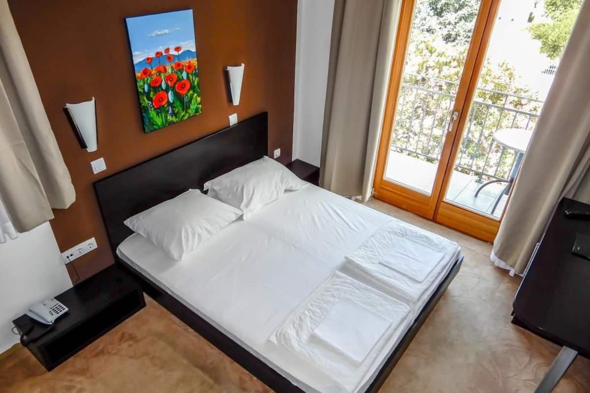 A neatly made bed with white linens, a painting of red flowers on the wall, and a view of greenery through the window.