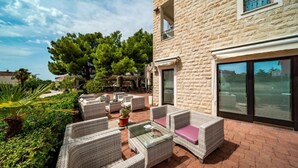 Un patio con muebles de mimbre y una mesa de vidrio, rodeado de follaje y un edificio de piedra.