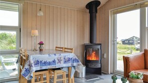 A cozy room with a wooden interior, a table with chairs, a table lamp, a vase with flowers, a fireplace, and a view of the outside. - Cozy home in Hvide Sande with kitchen (Hvide Sande)