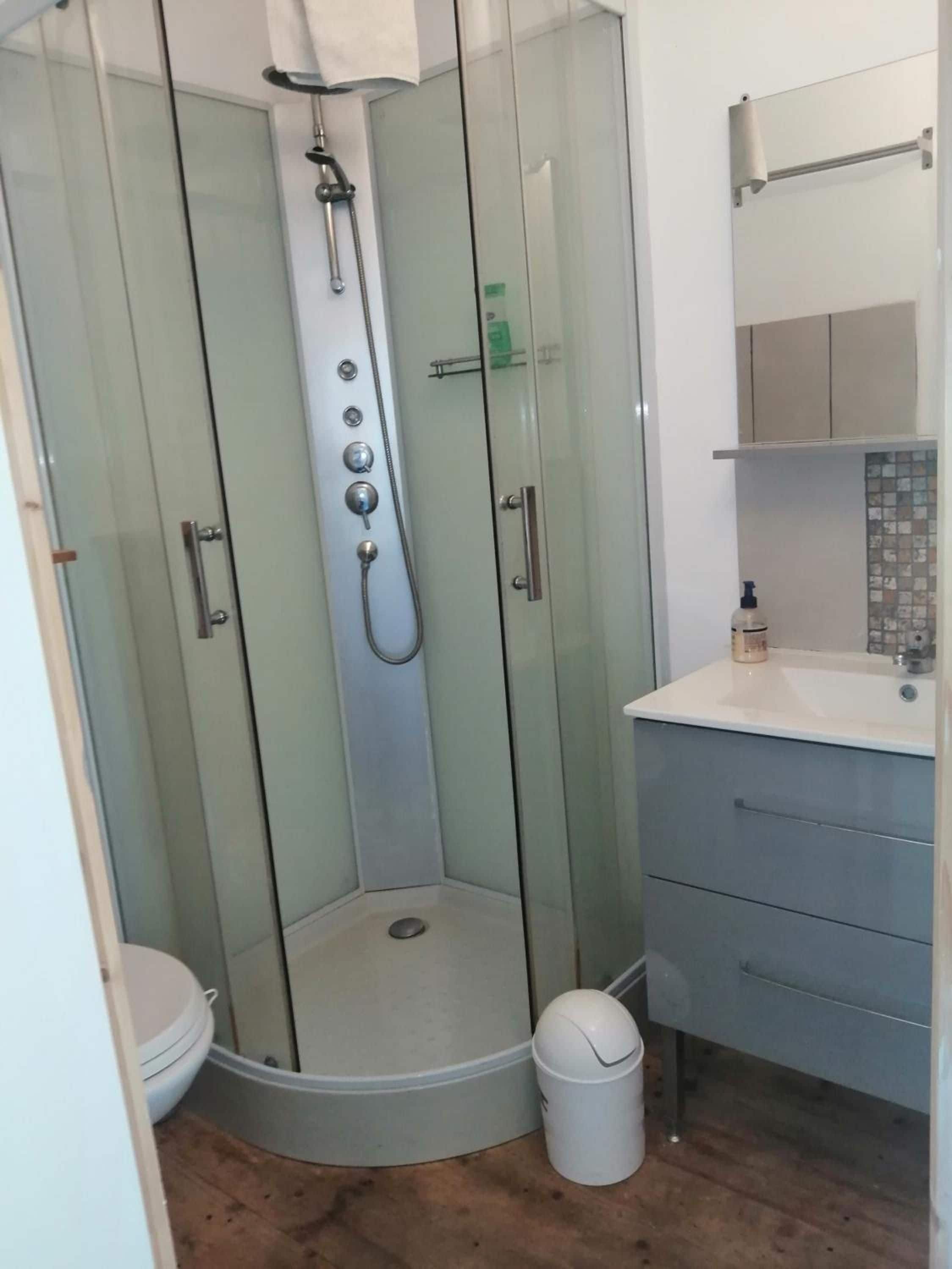 Cabina de ducha con paredes de vidrio, cabezal de ducha de mano, inodoro y lavabo con espejo.