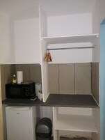 Una pequeña cocina americana con microondas, un refrigerador compacto, un estante con objetos colgando y un bote de basura.