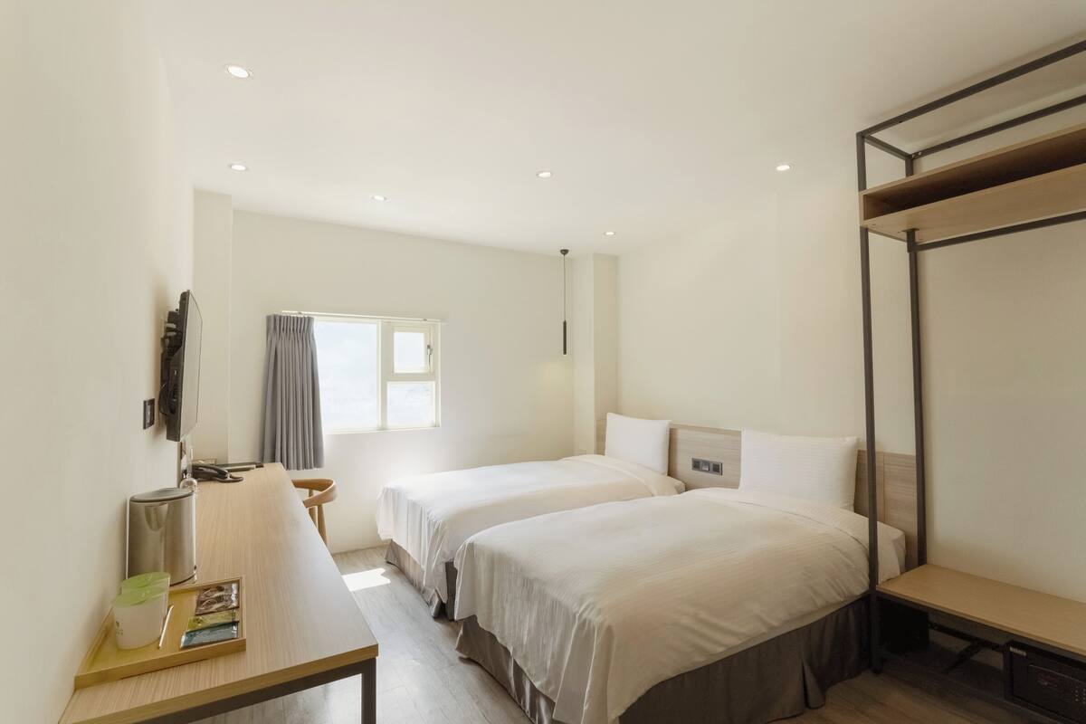 Ein Hotelzimmer mit zwei Betten, einem Nachttisch, einem Schreibtisch, einem Stuhl, einem Fernseher, einem Fenster mit Vorhängen und einer Deckenleuchte.