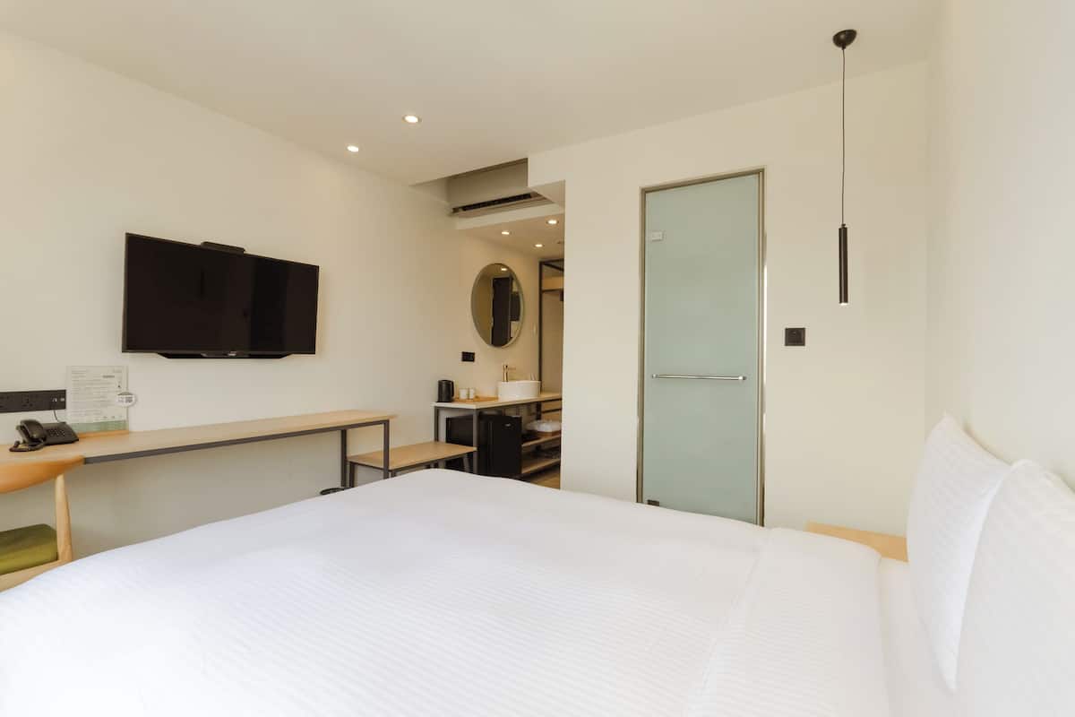 Ein Hotelzimmer mit einem großen Bett, einem Schreibtisch mit Telefon, einem flachbildfernseher an der Wand, einer kleinen Küchenzeile und einer Tür zu einem Badezimmer.