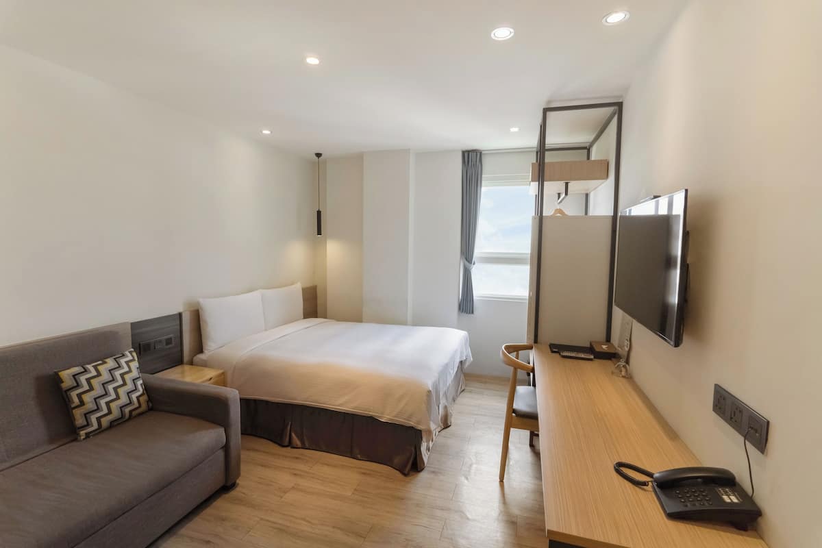 Ein modernes Hotelzimmer mit einem Bett, einer Couch, einem Schreibtisch mit Telefon, einem Fernseher und einem Fenster mit Vorhängen.