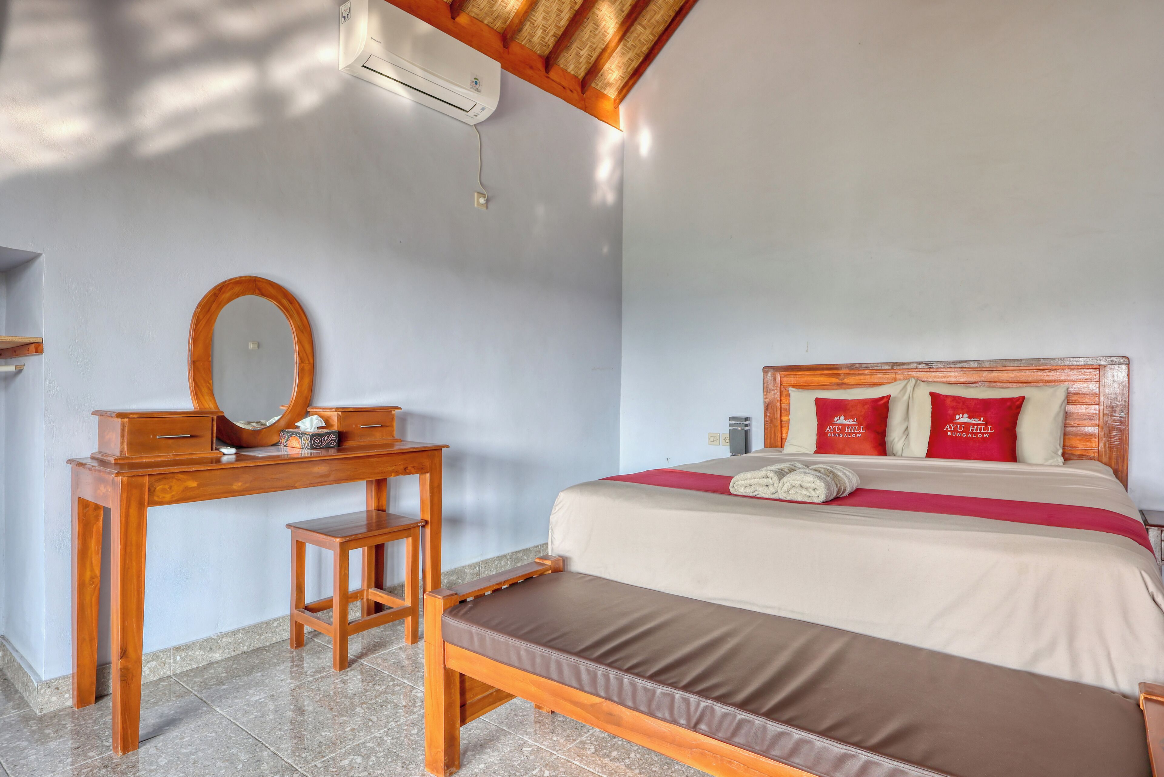 A bedroom with a bed, a wooden desk with a mirror, a bench, and a wall-mounted air conditioner.