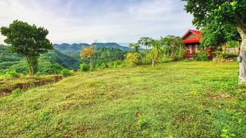 A grassy area with a small building with a red roof, surrounded by trees and hills in the background.