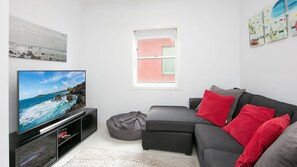 En moderne stue med et fladskærms-tv, en grå sofa, røde og grå puder, en lille poselejemøbel og et vindue med persienner.