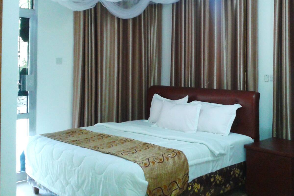 A neatly made bed with white linens and a decorative throw, a wooden nightstand beside it, and curtains with a striped pattern.