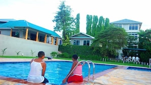 Zona de piscina con dos personas sentadas al borde, rodeada de edificios y vegetación.