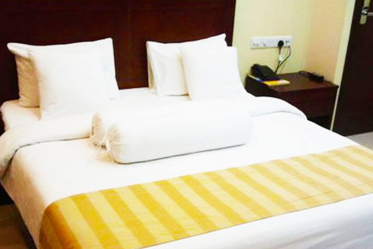 A hotel room with a neatly made bed, a bedside table with a phone and a lamp, and a door to the right.