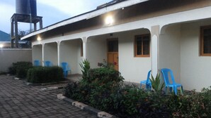 A building with a covered patio area, blue plastic chairs, and potted plants. - Hotel Premier (Yumbe)