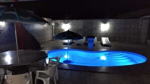 Una piscina exterior amb llum blava, envoltada de cadires i una taula de plàstic blanc.