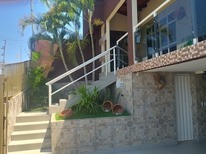A residential building with a staircase leading to a balcony, surrounded by palm trees and potted plants.