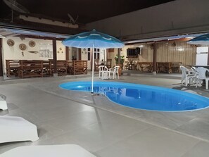 A swimming pool area with a blue pool, surrounded by white plastic chairs and tables, and a ceiling fan.