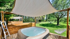 Un jacuzzi en una terraza de madera con toldo, rodeado de césped y árboles.