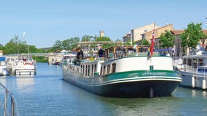 Un barco de recreo amarrado en un muelle, junto a otros barcos, con un puente al fondo y edificios a lo largo de la orilla del río.