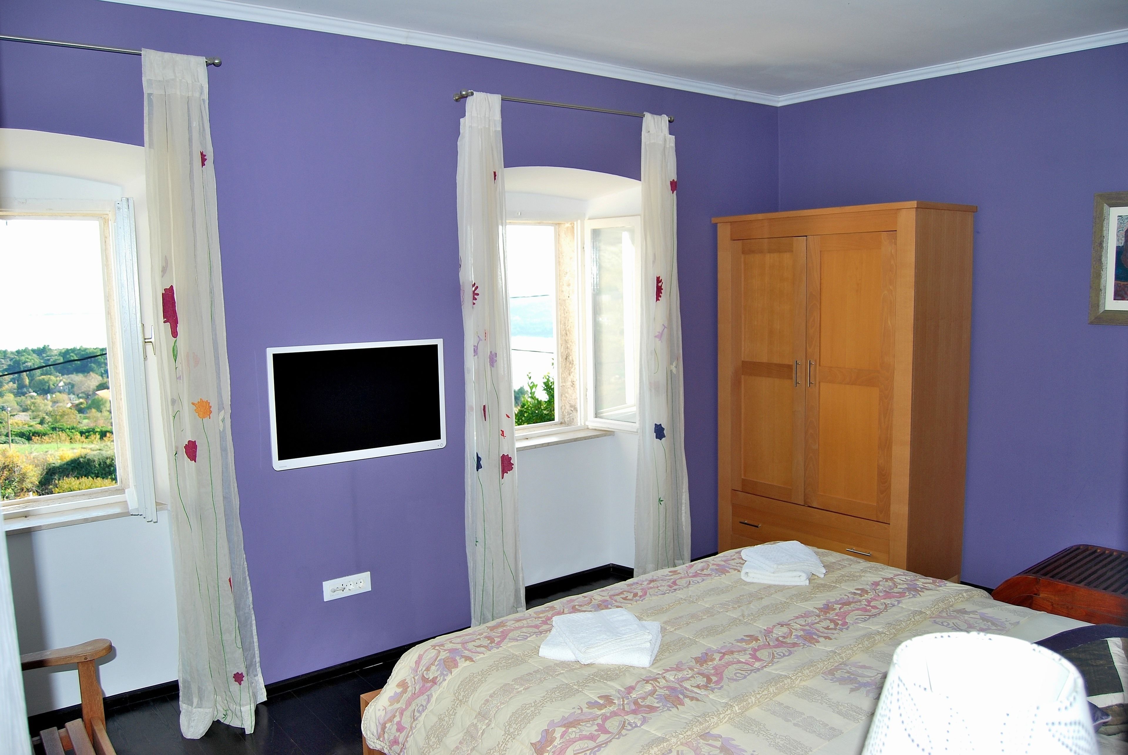 A bedroom with purple walls, a bed with a floral bedspread, a wooden wardrobe, a television on the wall, and a window with curtains.