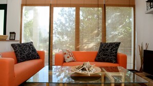 A living room with two orange sofas, a glass coffee table, and a large window with blinds. - Elies Villa (Kalymnos)
