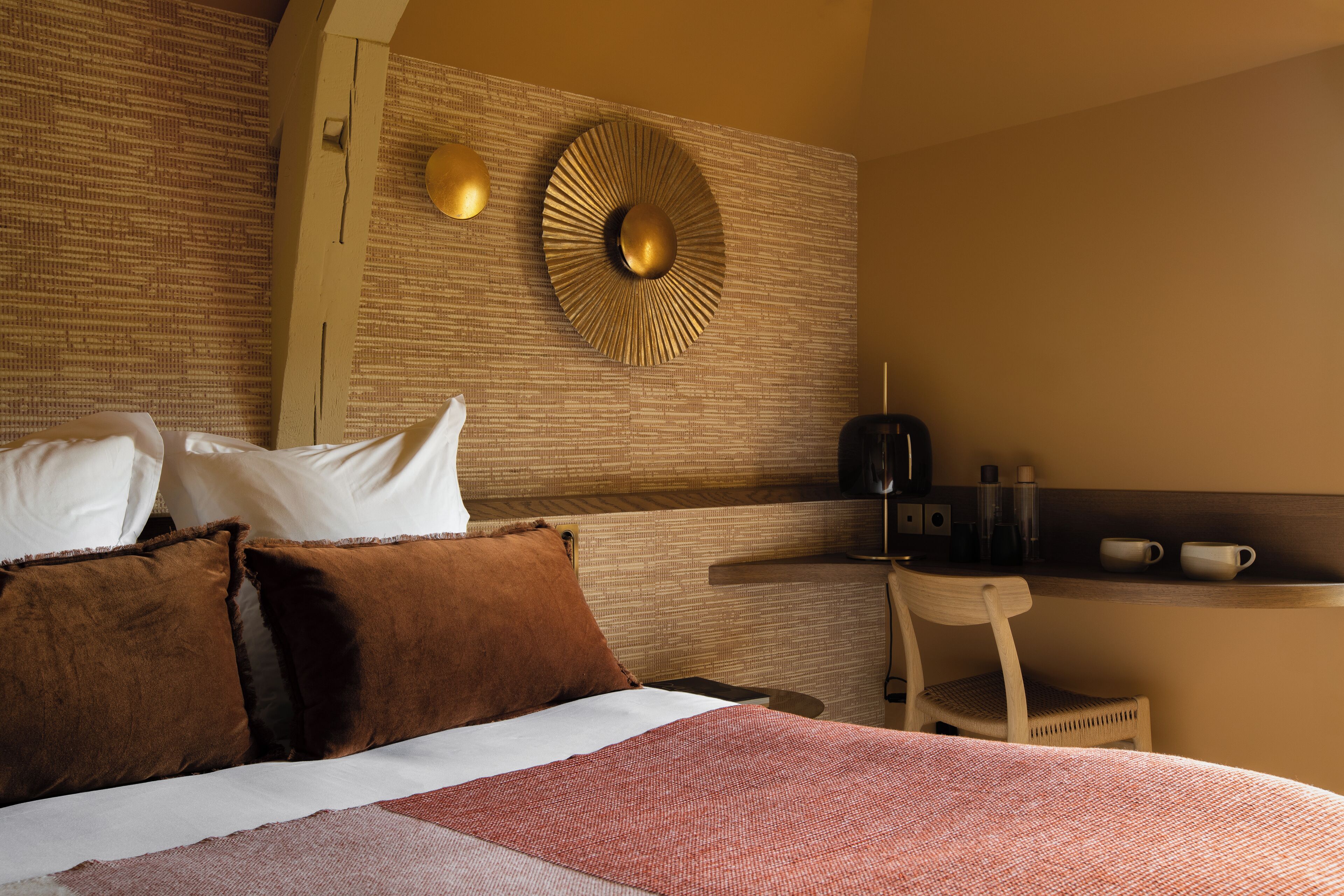 Habitación de hotel con cama, almohadas, un adorno circular en la pared, una mesita con tazas y una silla.
