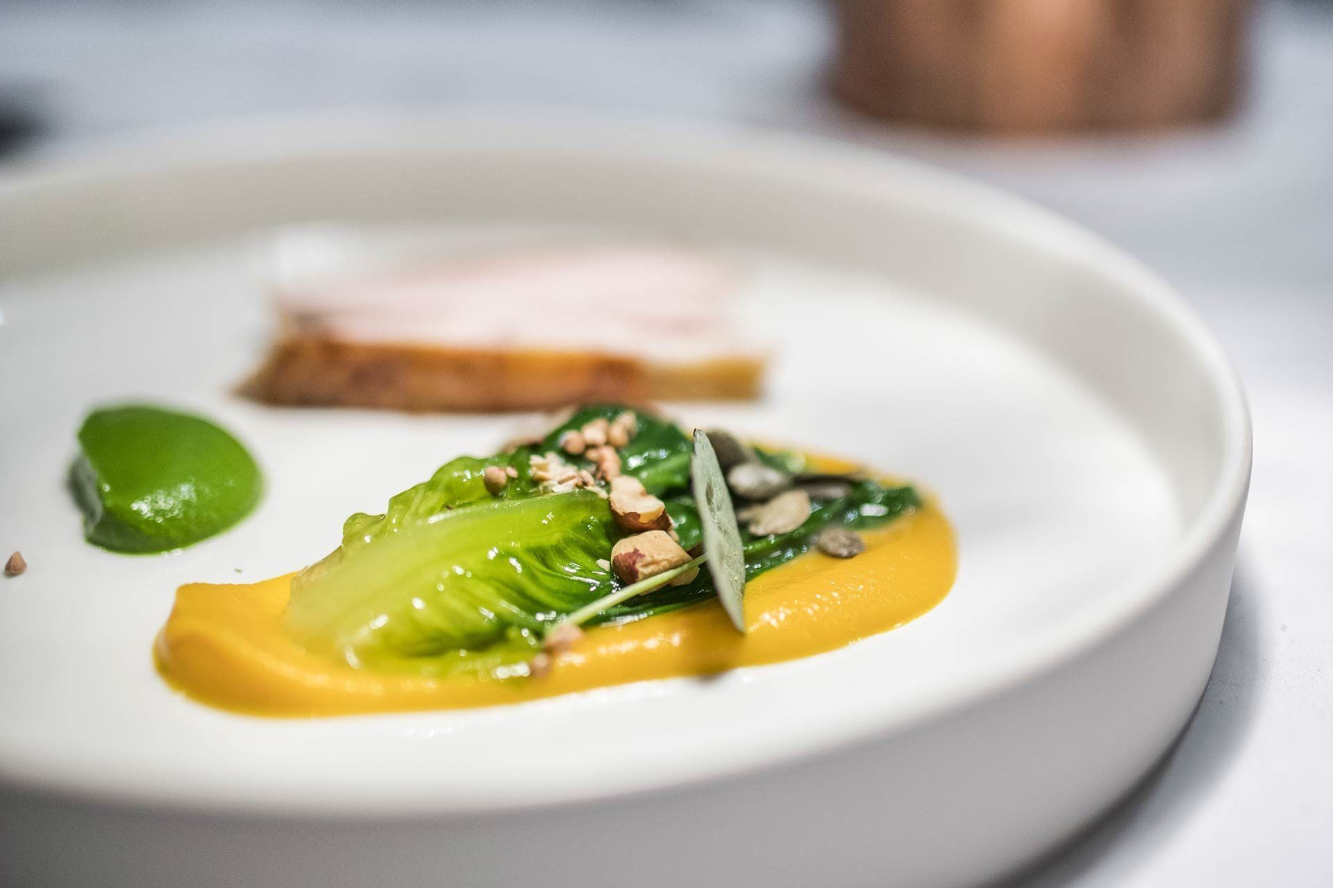 A plate with a dish of greens, nuts, and a yellow sauce, and a slice of meat on a white plate.