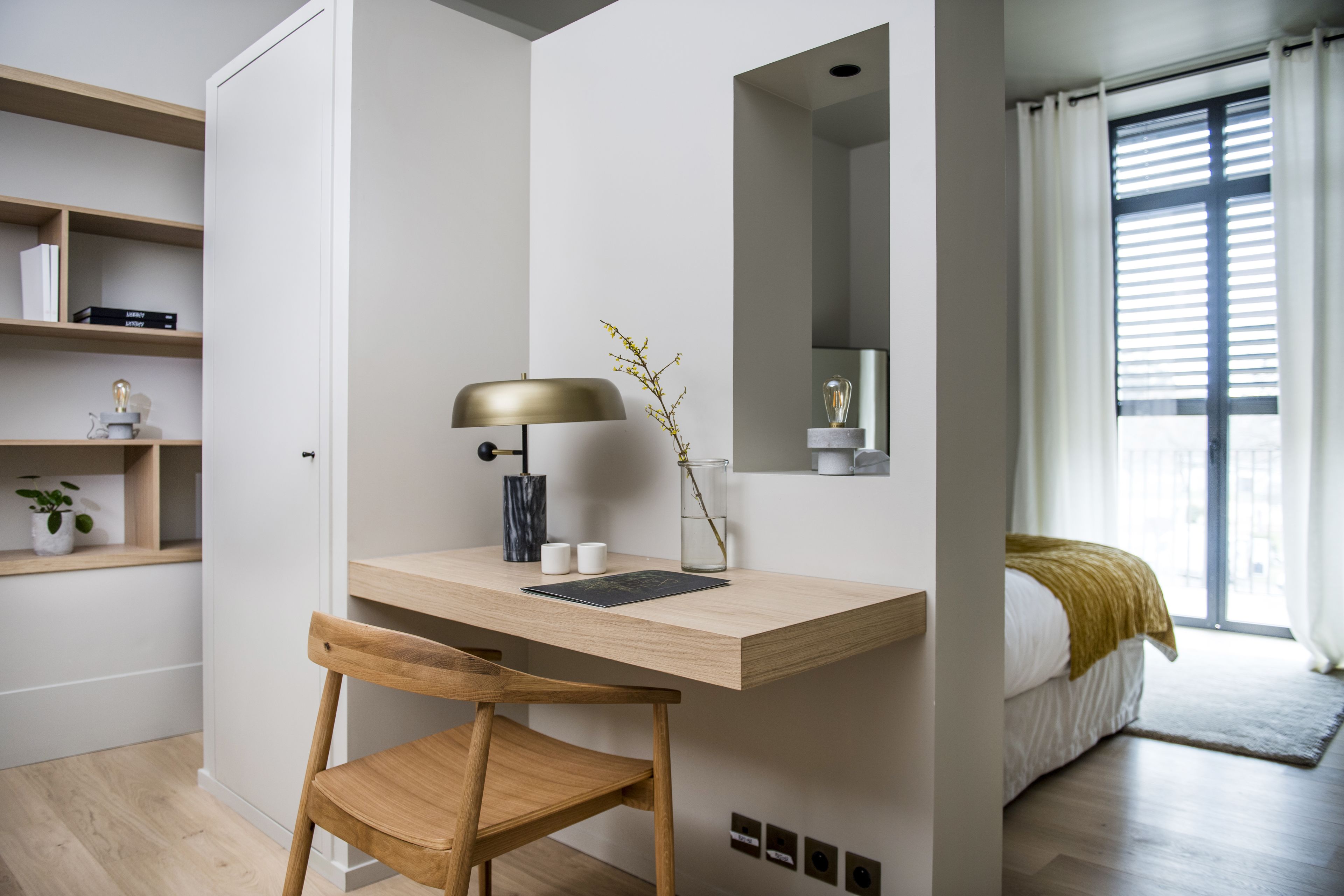 Ein modernes Schlafzimmer mit einem Holztisch, einem Stuhl, einem Nachttisch, einer Lampe, einem Spiegel und einem Fenster mit Vorhängen.