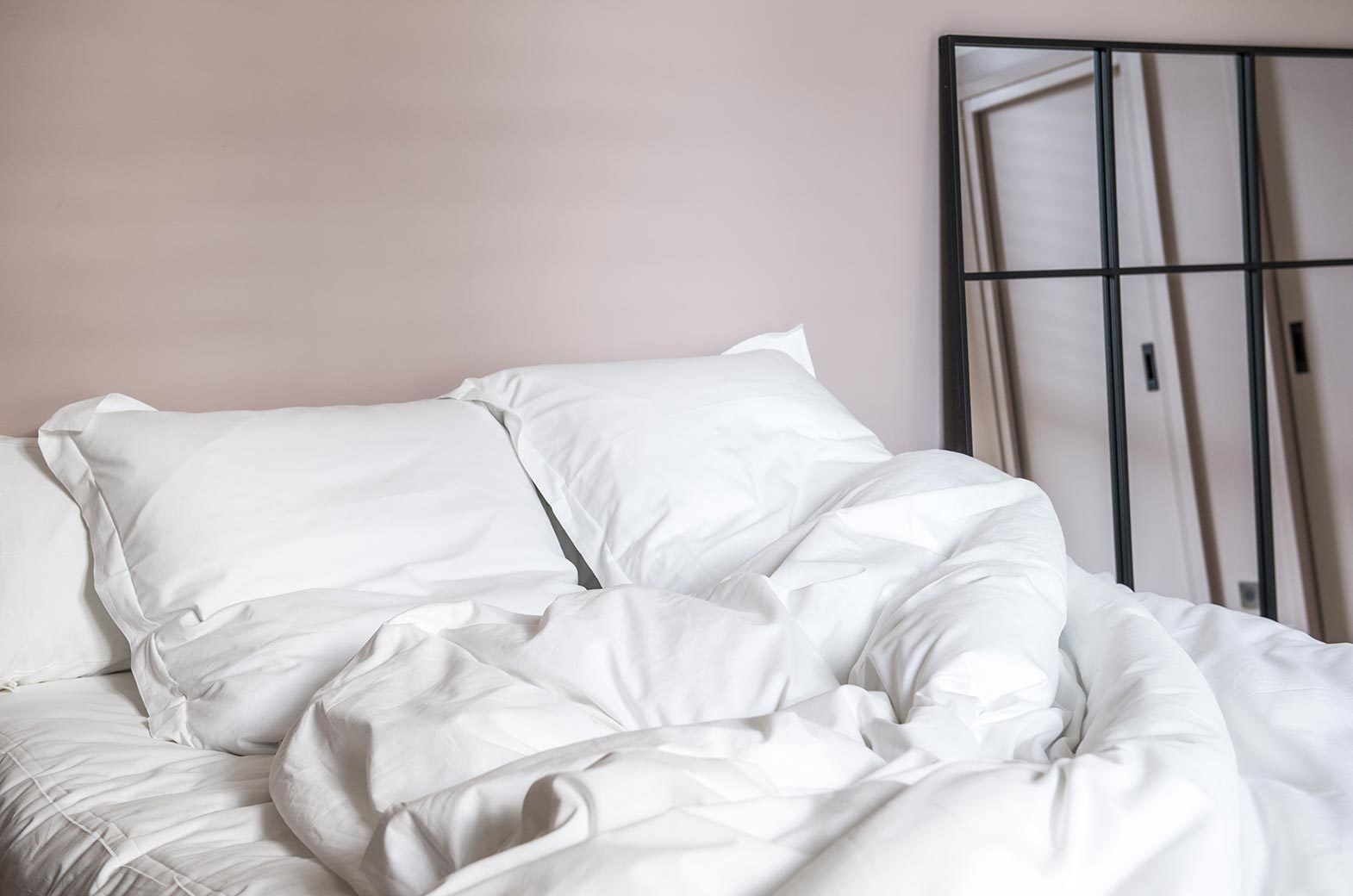 A bed with white bedding in a room with a plain wall and a large window.