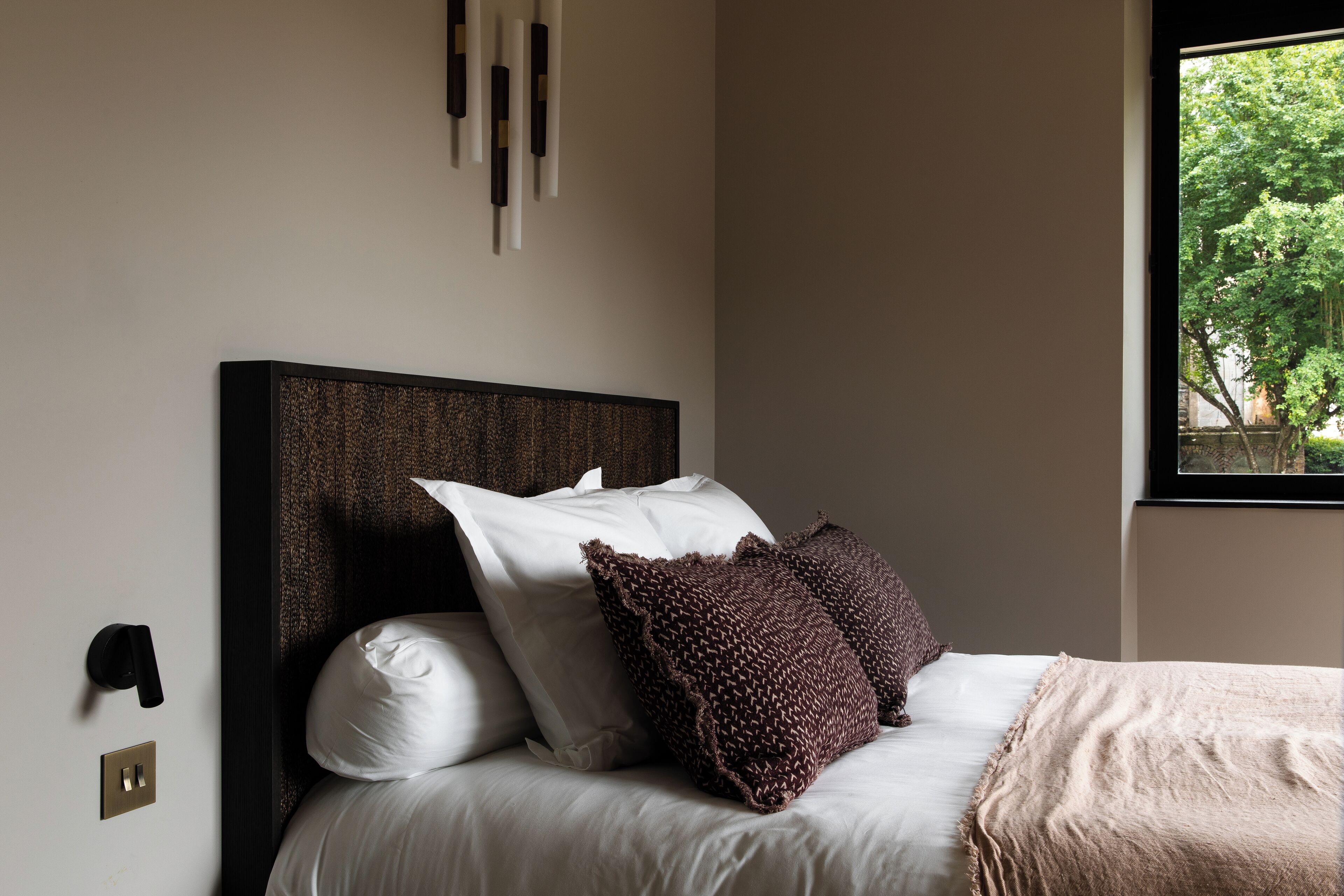 A neatly made bed with white linens and a brown throw blanket, a wall-mounted light fixture, and a window with a view of trees.
