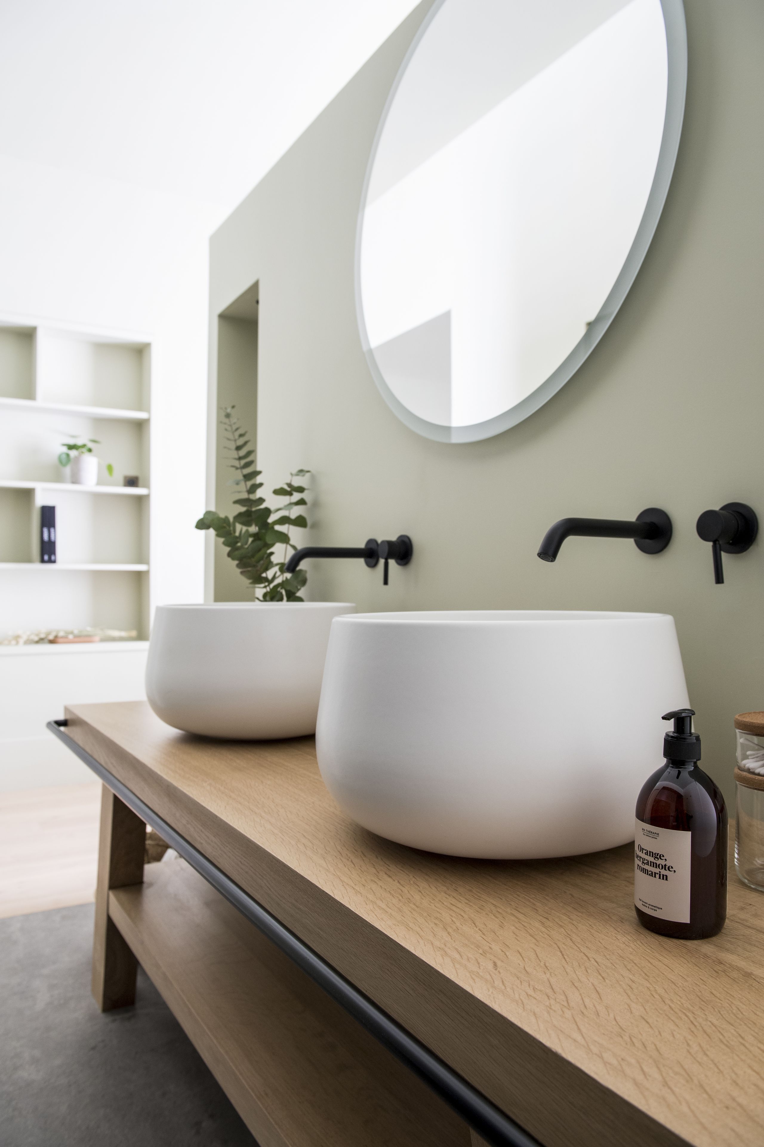 A modern bathroom with a wooden vanity, two large white sinks, a round mirror, and a black faucet.