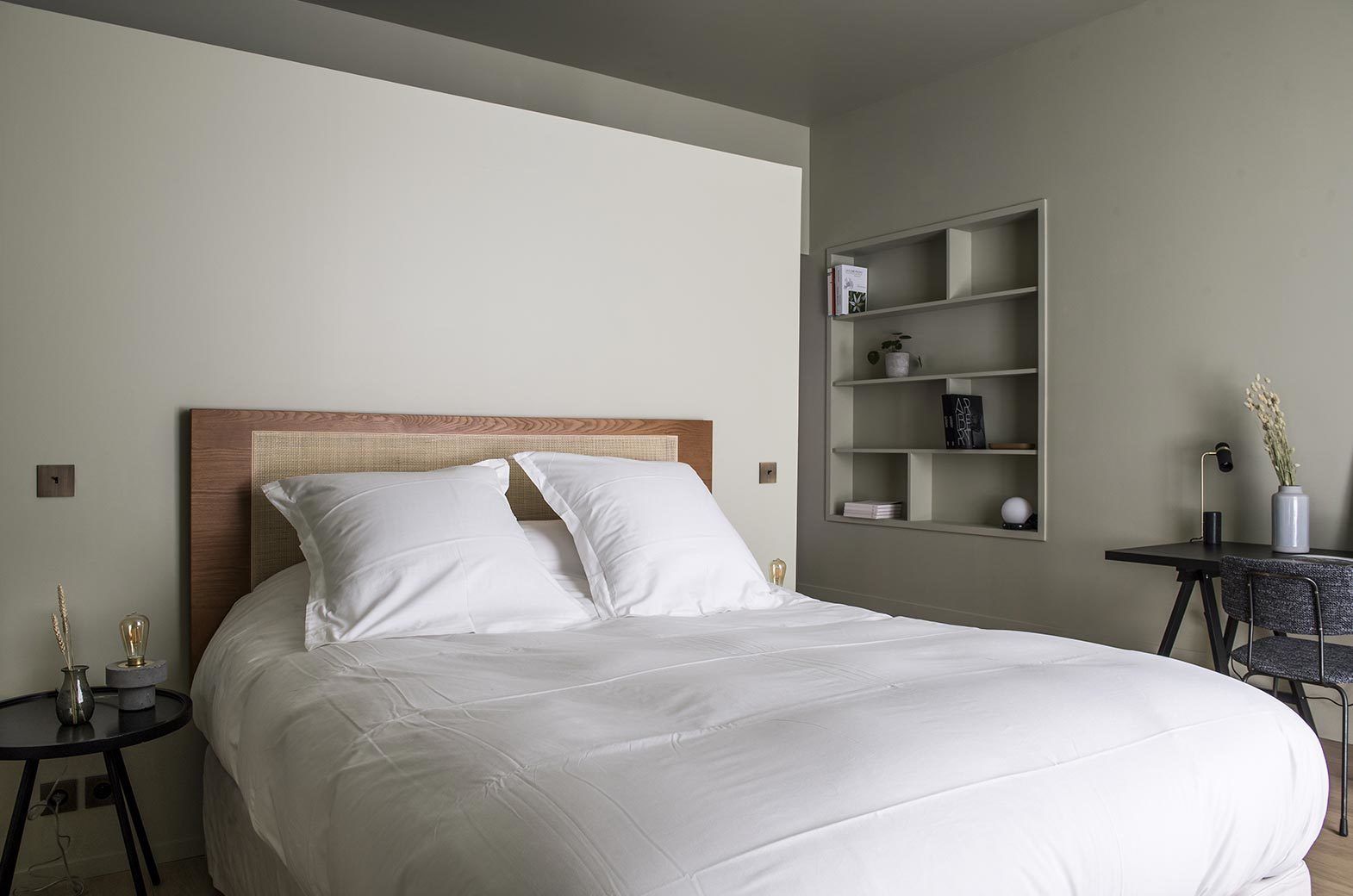 A neatly made bed with white linens and a wooden headboard, a small bedside table with a lamp, and a wall-mounted shelf with books and decorative items.