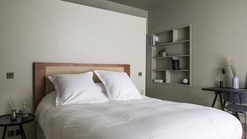 A neatly made bed with white linens and a wooden headboard, a small bedside table with a lamp, and a wall-mounted shelf with books and decorative items.