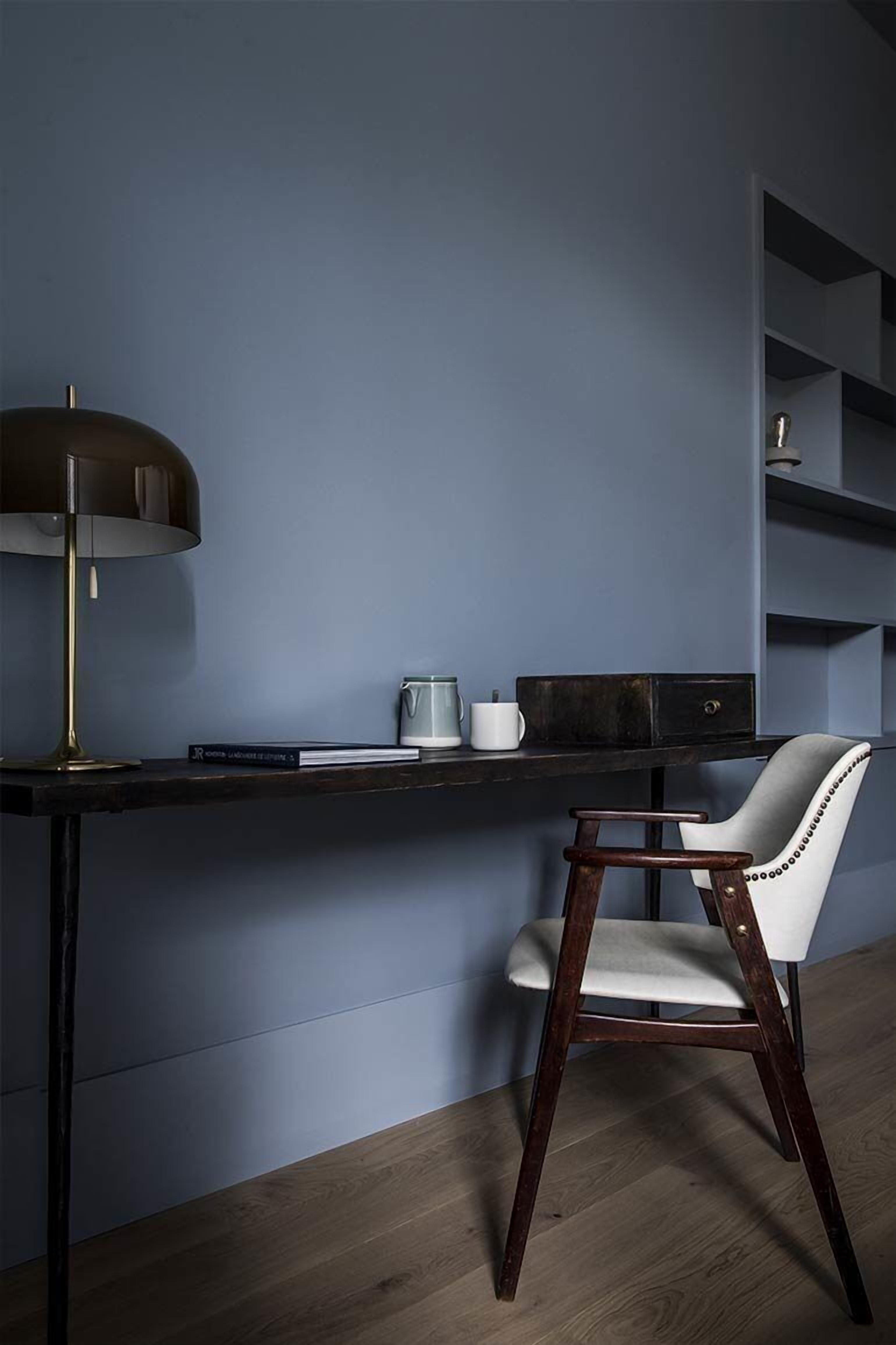 Un escritorio con lámpara, una silla y una estantería frente a una pared azul.