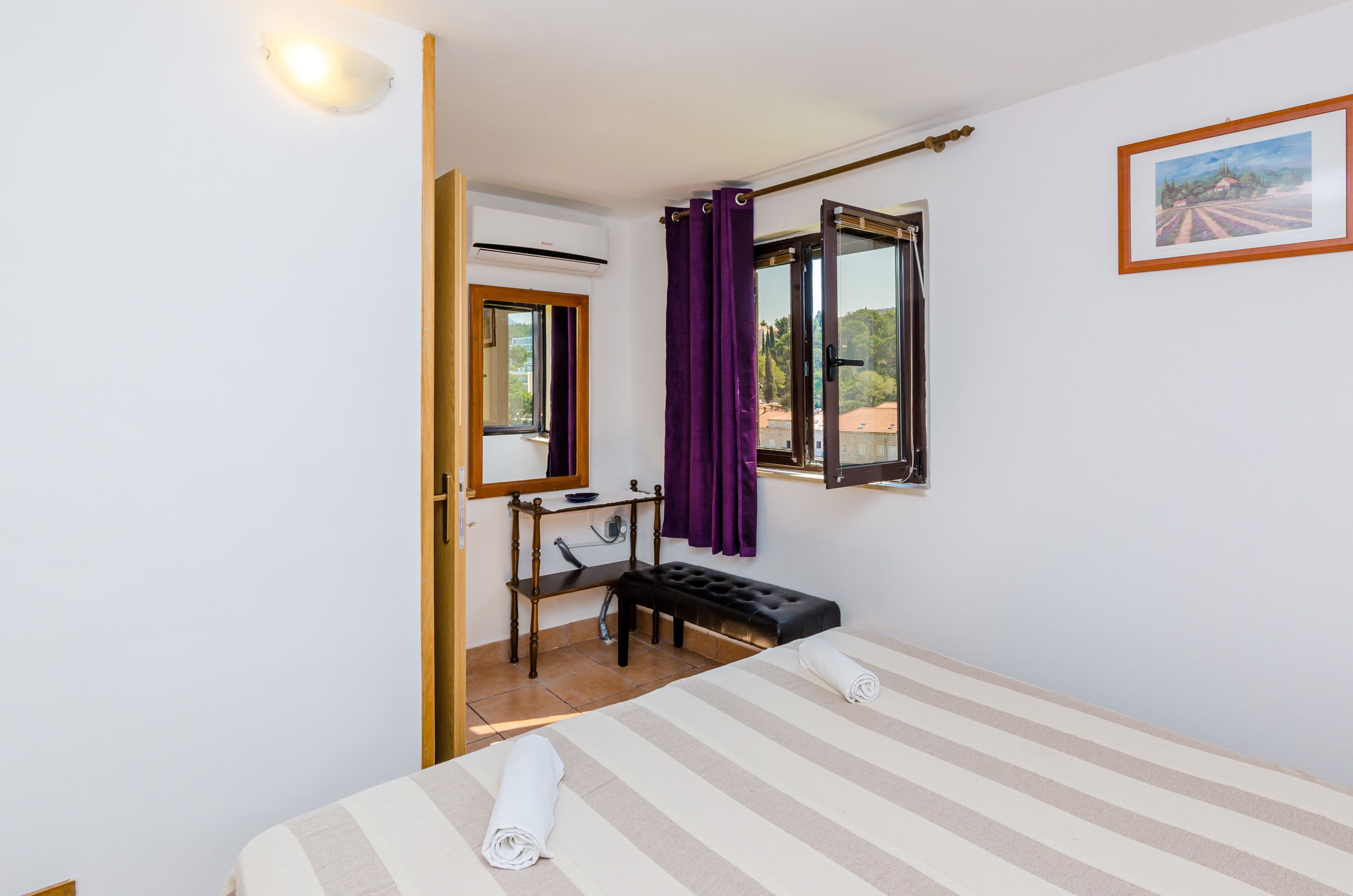 Hotellihuoneessa on sänky, penkki, peili, ikkuna, jossa on violetit verhot, ja seinällä on kehystetty taulu.