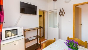 Une petite pièce équipée d’un four à micro-ondes, d’une télévision, d’une chaise en bois, d’une table ornée d’un vase avec une fleur violette, et d’une porte donnant sur une autre pièce.