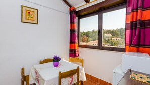 Un balcon avec une table et des chaises, une fenêtre avec des rideaux, une plante en pot et une vue sur des immeubles.