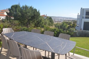 A rectangular outdoor table with a marble-like surface, surrounded by six chairs, set against a backdrop of greenery and buildings.