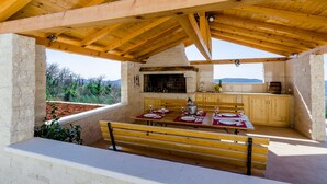 An outdoor kitchen with a stone fireplace, wooden beams, and a dining table set for four. - Villa Dragic (Konavle)