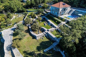 Vue aérienne d’une propriété comprenant une maison en pierre, une piscine et un jardin aménagé avec des allées et des arbres.