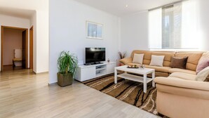 Een moderne woonkamer met een beige bank, een flatscreen televisie op een witte standaard, een salontafel en een groot raam met jaloezieën.