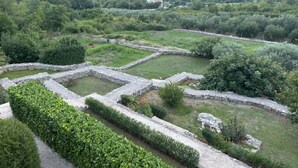 Een historisch plekje met stenen muren en paden, omgeven door weelderige begroeiing en een gebouw in de verte.