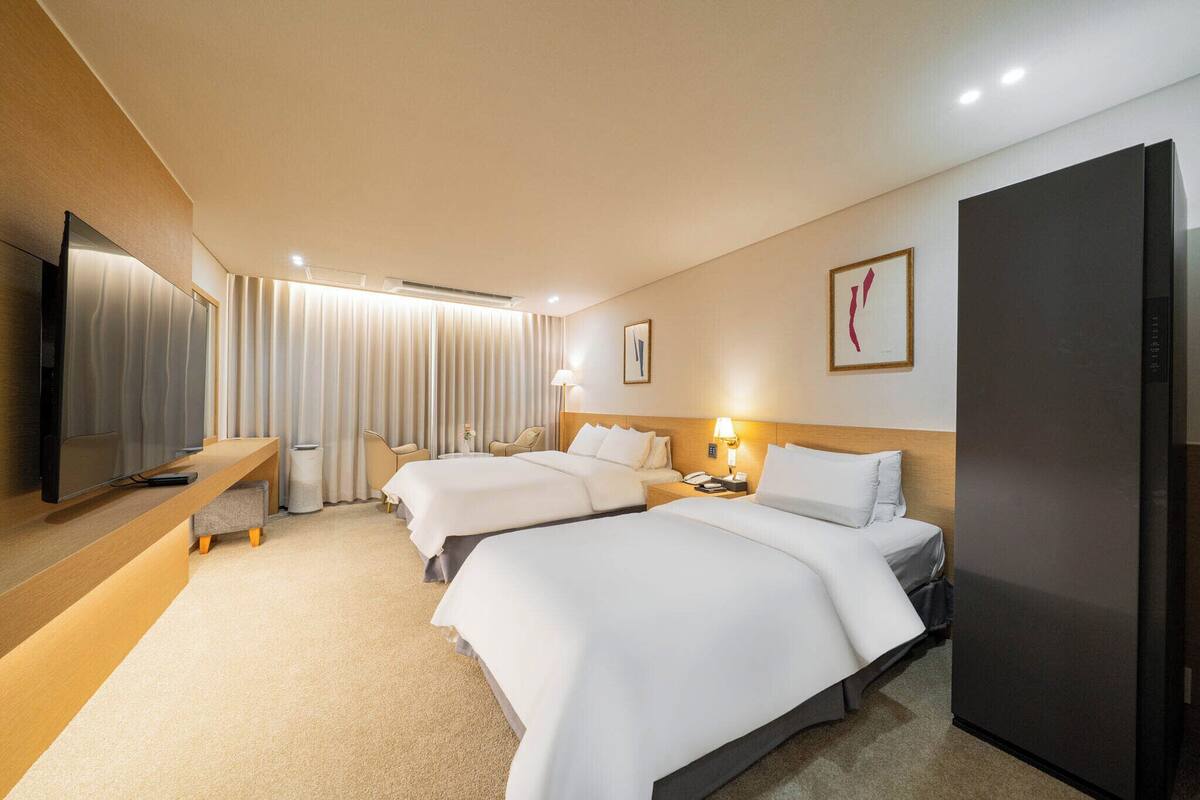 A hotel room with two beds, a TV, a desk, a refrigerator, and a bathroom with a shower.
