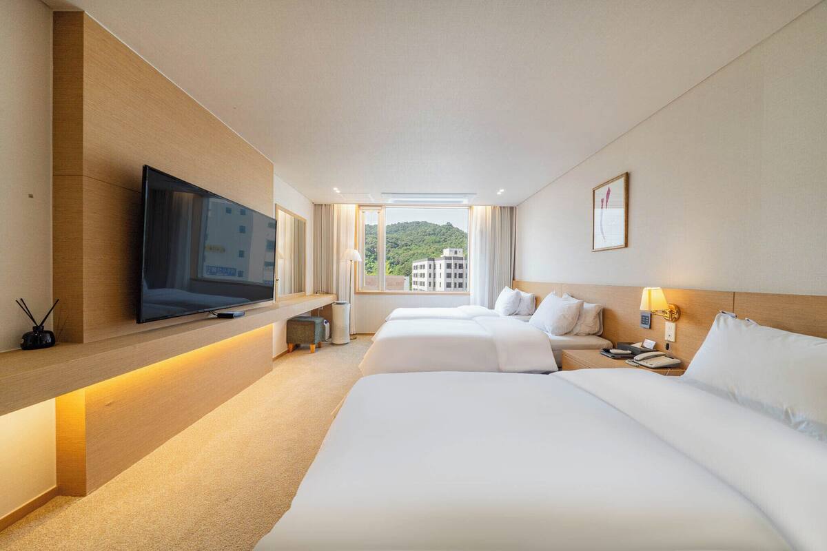 A hotel room with a TV on a wooden shelf, two beds, a bedside table with a lamp, and a window with a view of buildings.