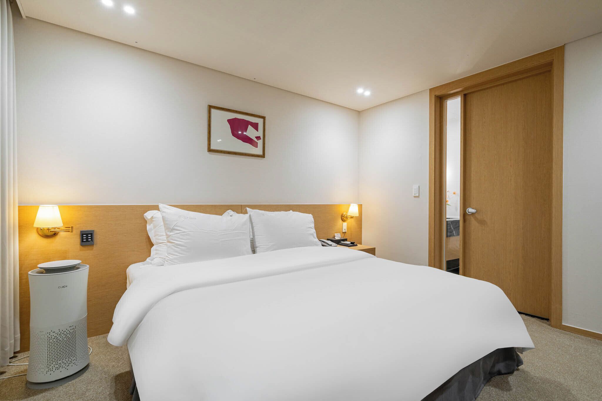 Habitación de hotel con una cama, dos lámparas de noche, un cuadro en la pared, una puerta de madera y un calentador portátil.