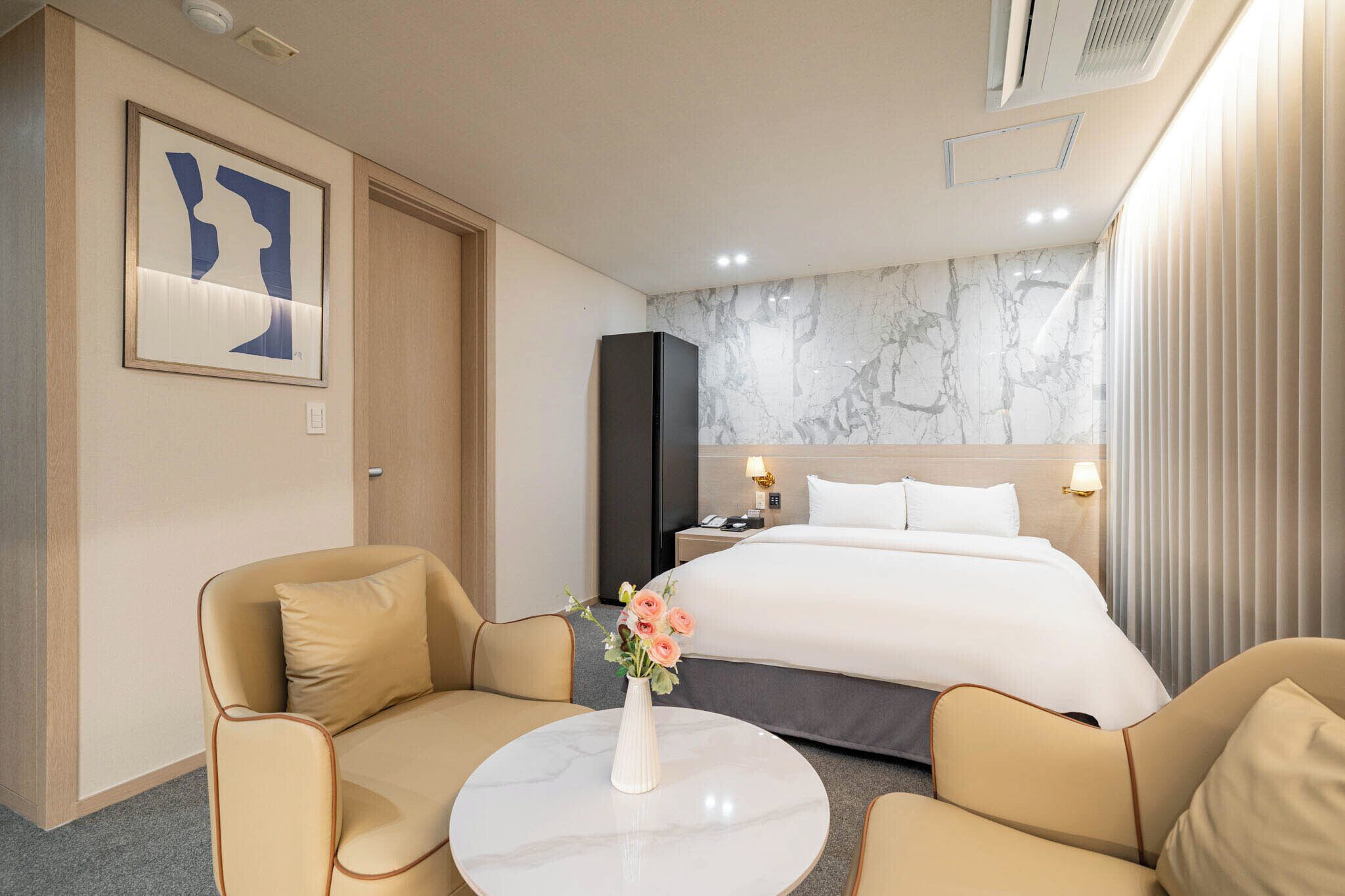 Habitación de hotel con cama, dos sillones, una mesita con un florero, pared de mármol y una obra de arte enmarcada.